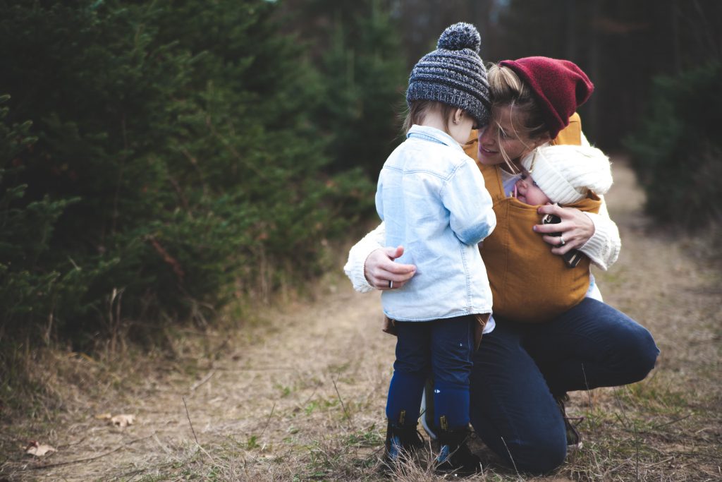 Woman and two children kneeling on a dirt path in nature, exploring together surrounded by greenery