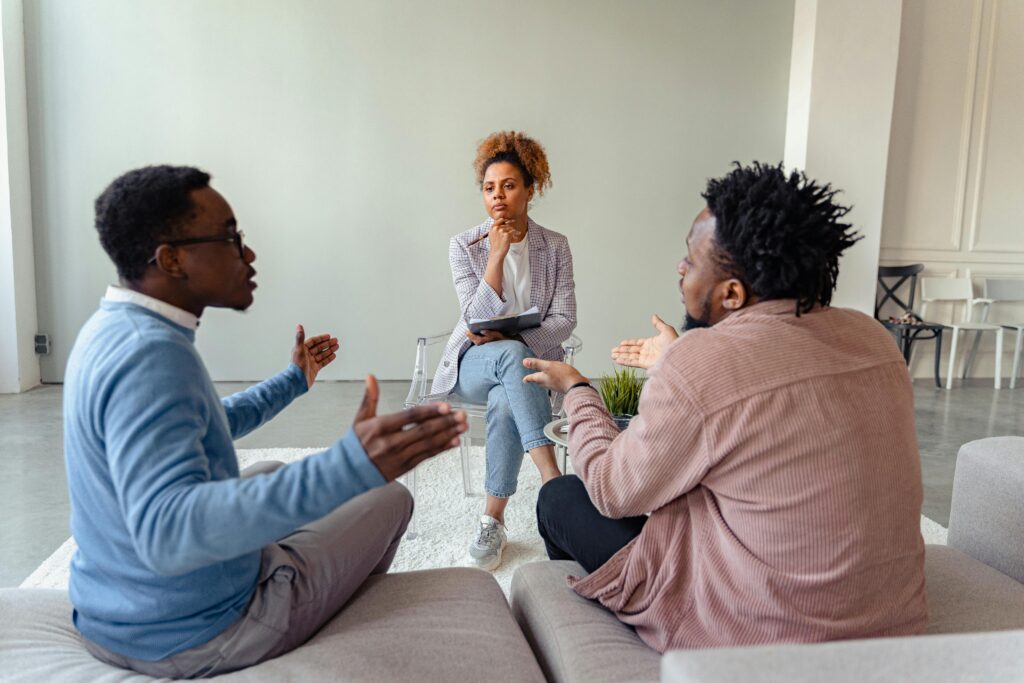 Female counselor talking with two men during a therapy session in a cozy living room